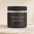 A jar of Nouvelle Eden Glow Révolution Revitalizing Body Scrub, featuring a black ribbed lid and a dark brown label with gold and white text, set against a neutral beige background.