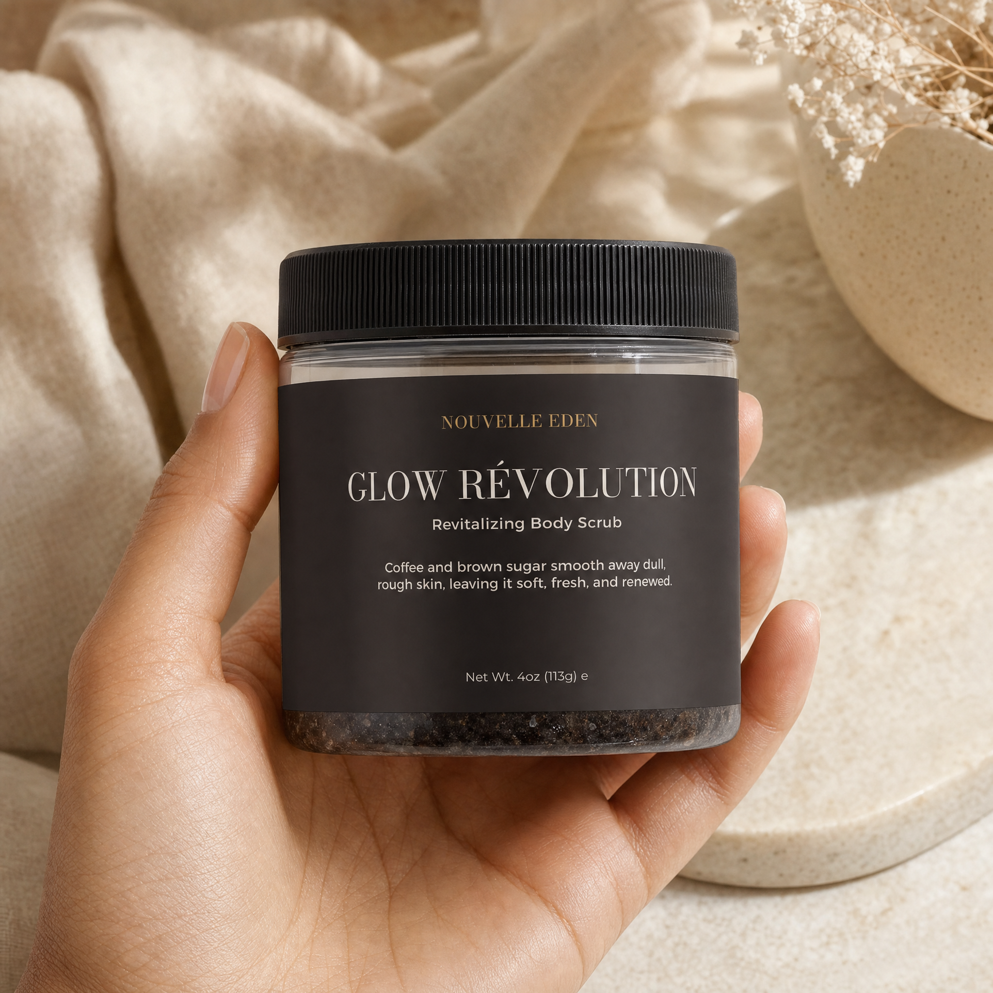 A hand holds a clear jar of Nouvelle Eden Glow Révolution Revitalizing Body Scrub, showing the black label and black lid. The background features a draped beige fabric and a ceramic dish.